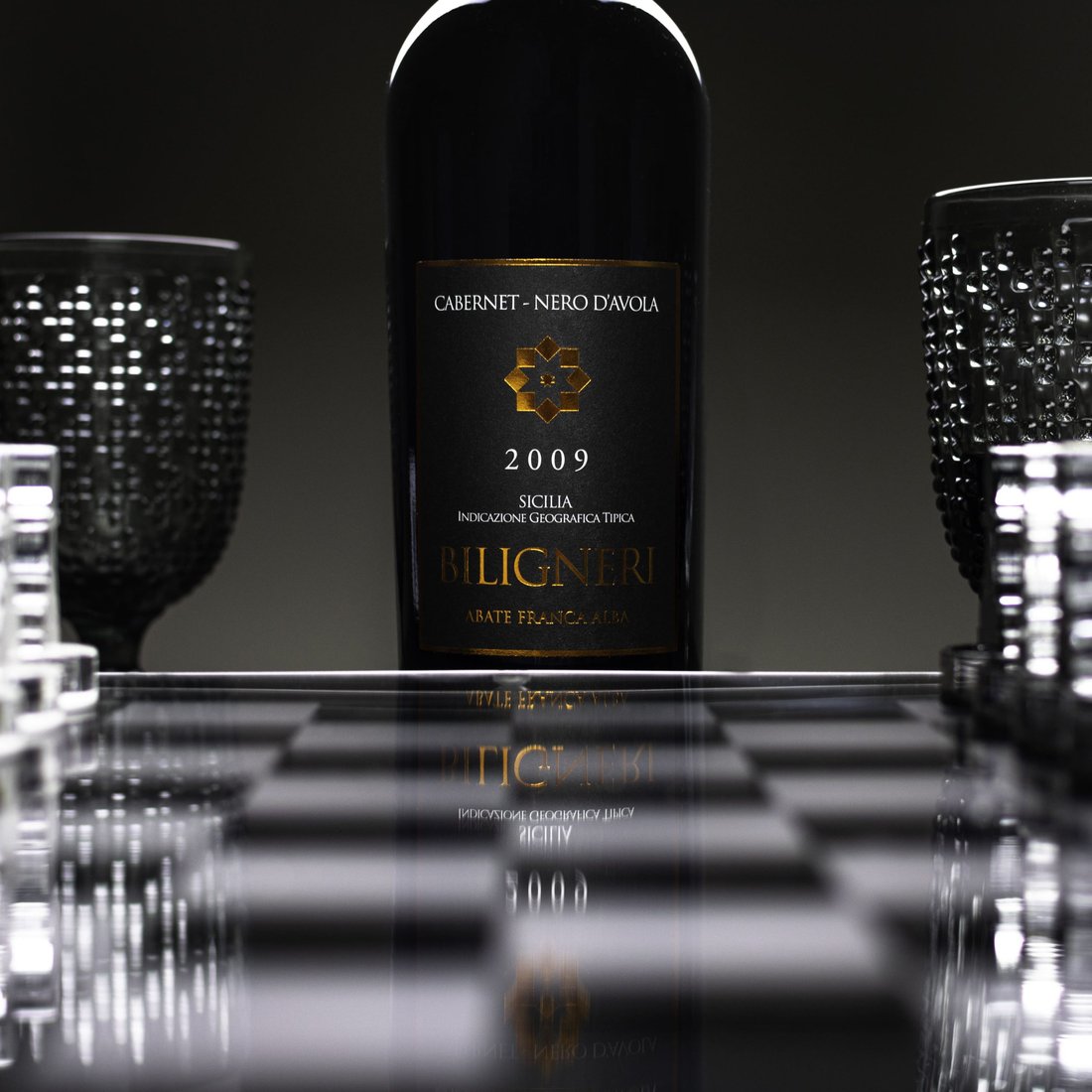 Bottiglia di Biligneri Cabernet-Nero d'Avola 2009 Abate Franca Alba tra calici scuri a rilievo e scacchiera in vetro
